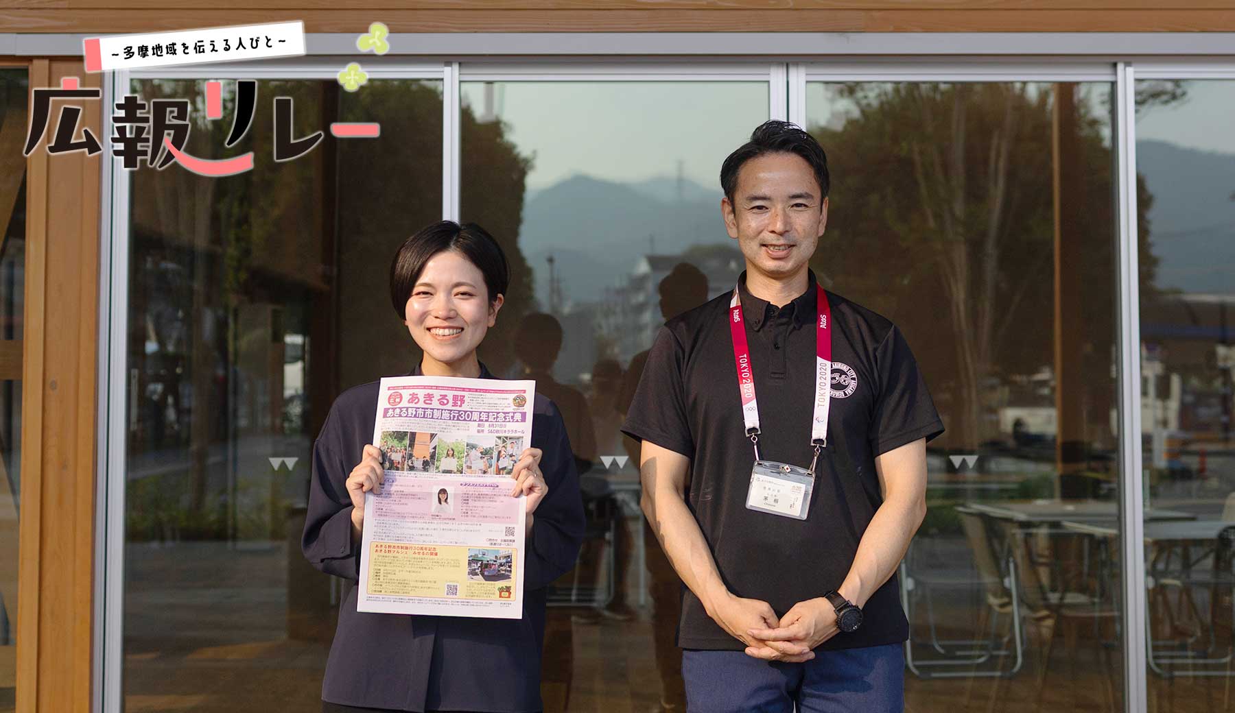 インタビューを行ったフレア五日市の前に立つあきる野市広報担当の豊島さん（左）と茅根さん（右）。豊島さんはあきる野市の広報紙を持っている。（左上に多摩地域を伝える人びと 広報リレー のロゴ入り）