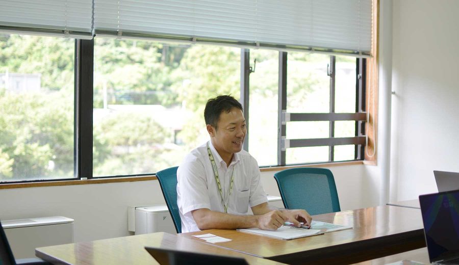 奥多摩町役場の会議室で、大きな窓いっぱいに見える山々の緑を背に、インタビューに応じる森田さん。