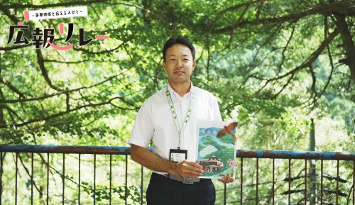 白髭神社境内の木々を背に、奥多摩町 町制70周年記念紙を持った広報担当の森田さんの写真。