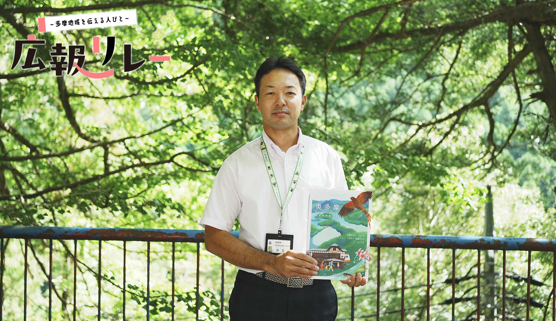 白髭神社境内の木々を背に、奥多摩町 町制70周年記念紙を持った広報担当の森田さんの写真。
