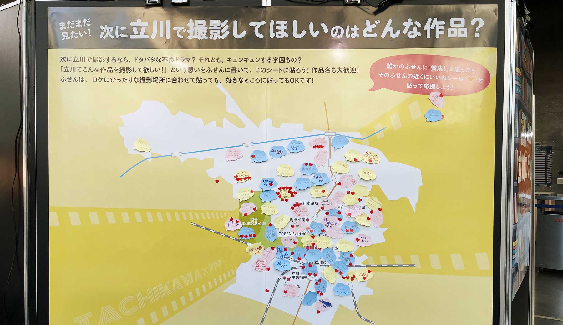 立川ロケーションサービス presents ロケ地の街 立川展 2026 会場内来場者参加型企画のパネル。次に立川市で撮影して欲しいのはどんな作品？と題して、来場者が撮影して欲しい映画のジャンルやタイトルなどを付箋に書いて貼り付けている。誰かの意見に賛同する人は、ハート型シールを貼って応援することもできる。